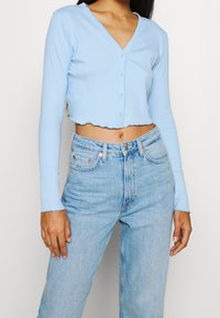 Vaaleansininen ribbikuvioinen pitkähihainen crop-toppi, jossa on V-pääntie ja napillinen kiinnitys. Siinä on viimeistelemätön helma ja vartalonmyötäinen leikkaus, yhdistettynä vaaleansinisiin farkkuihin.