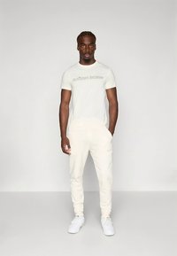 Svetlý set športového oblečenia pozostávajúci z jednofarebného trička a teplákov. Materiál je hladký; obe časti majú jemné prvky s logom značky.