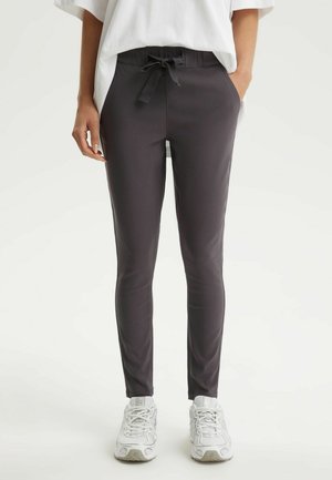 Osoba ubrana w czarne spodnie dresowe ze ściągaczem, białe trampki i luźną białą bluzkę, stojąca z jedną ręką w kieszeni.