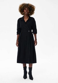 Czarna sukienka o kroju kopertowym z kołnierzykiem, trzema czwartej długości rękawami i akcentem w postaci guzika. Charakteryzuje się długością do midi oraz plisowanym wzorem. Zestawiona z czarnymi botkami za kostkę.