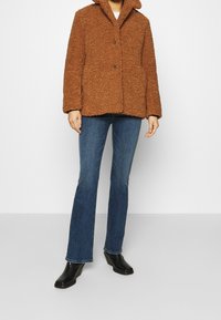 Brun texturerad fleecejacka med krage, framknappar och två sidofickor, kombinerad med utsvängda blå jeans och svarta ankelboots.