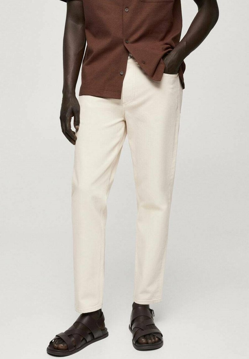 Pantaloni beige chiaro con vestibilità dritta, abbinati a una camicia a maniche corte marrone con bottoni e sandali marroni scamosciati.