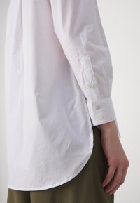 Lato destro di una persona che indossa una camicia bianca con maniche lunghe e polsini abbottonati, insieme a pantaloni verde oliva, su uno sfondo neutro.