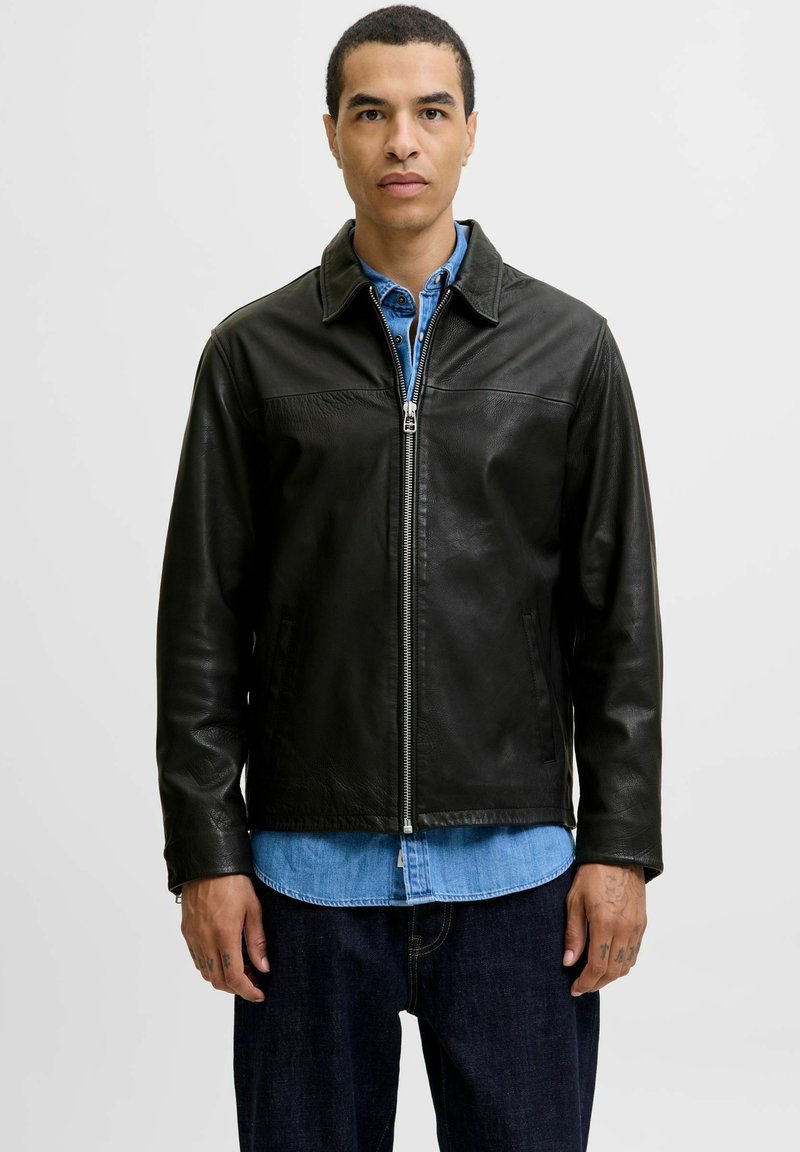 Black leather jacket with a collared neckline, front zip closure, and side pockets, worn over a denim shirt. Smooth texture and fitted design.