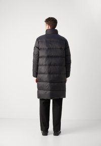 Schwarze Daunenjacke mit horizontalen Steppnähten, hohem Kragen und normalem Schnitt. Glattes, glänzendes Material; knielanges Design. Über schwarzen Hosen getragen.