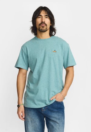 Man met halflang haar en baard, die een turquoise T-shirt draagt met een kleine auto- en vogelborduursel en blauwe spijkerbroek, staand met één hand in de zak.