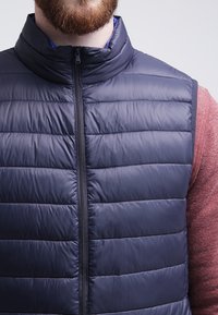 Námořnická puffer vesta s prošívaným vzorem, vysokým límcem a zipovým uzávěrem. Nosená přes strukturální vínovou košili s dlouhým rukávem.