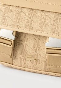 Μπεζ τσάντα με υφή και γεωμετρικό μοτίβο, μεταλλικά κούμπωματα, υφασμάτινα λουριά και μικρό λογότυπο κροκόδειλου στο κέντρο.