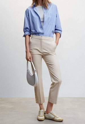 Frau trägt beige kurze Hosen mit Knopfdetails, blau gestreiftes Hemd über grauem Oberteil, beigen Sneakers und hält eine graue Handtasche.