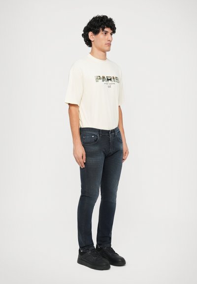 Ljuskrämsfärgad t-shirt med "PARIS" grafik i gröna nyanser, kombinerad med mörka jeans och svarta sneakers. Modellen står sidledes mot en vit bakgrund.
