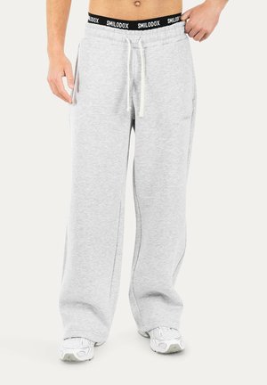 Personne portant un pantalon de survêtement ample gris clair avec une ceinture noire "SMILODOX" et des baskets blanches, tenant la ceinture d'une main.