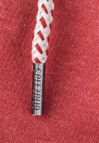 Rode stof met een witte en rode gevlochten trekkoord, met een metalen eindstuk gegraveerd met "SPITZBUB." Textuur zichtbaar in de gebreide achtergrond.