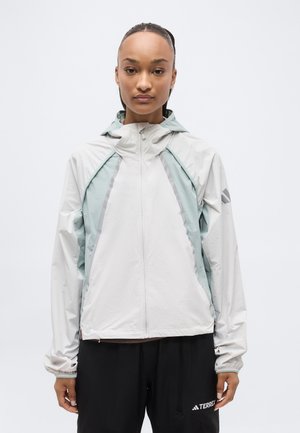 Frau trägt eine hellgraue und minzgrüne Kapuzenjacke mit Reißverschluss und schwarze Hose, steht vor einem einfachen weißen Hintergrund.