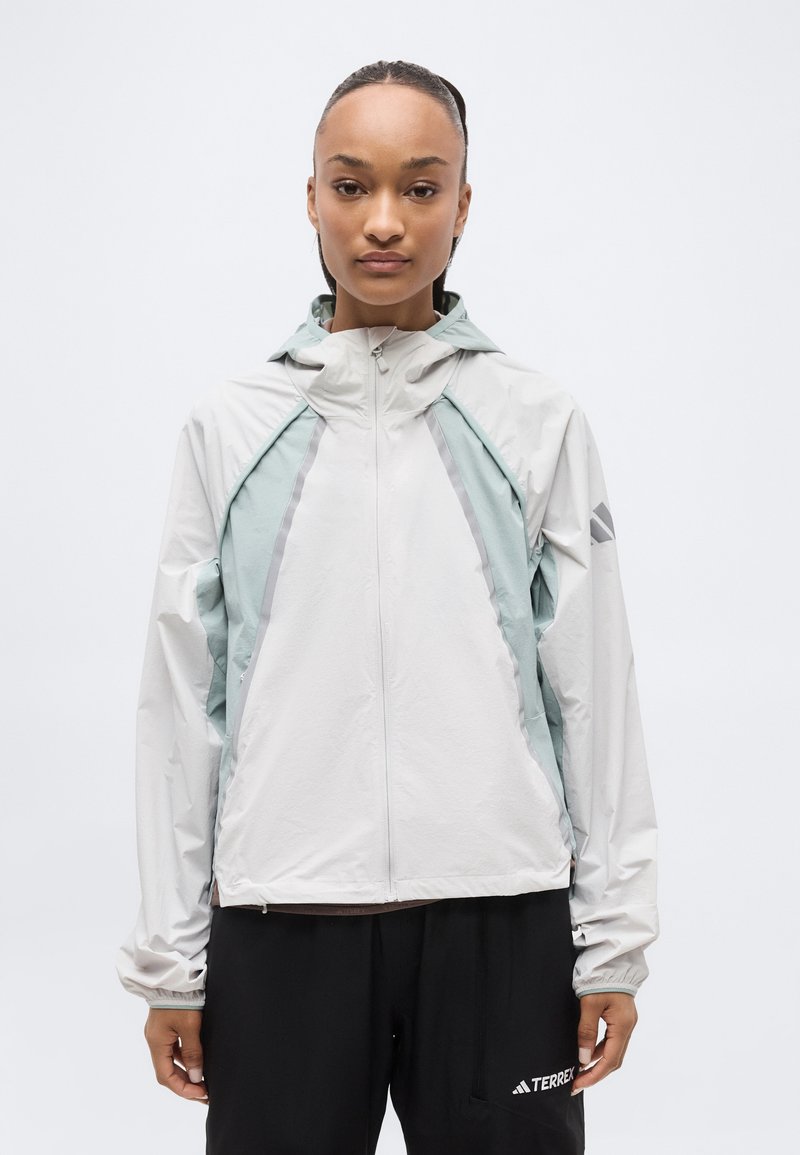 Γυναίκα που φοράει ελαφρώς γκρι και μέντα πράσινο μπουφάν με φερμουάρ και κουκούλα, μαζί με μαύρο παντελόνι, στέκεται μπροστά σε απλή λευκή φόντο.