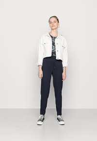 Jaqueta de ganga cropped branca, top escuro com padrão floral, calças navy com cordão e ténis pretos com solas brancas. Detalhes de hardware simples e textura lisa.