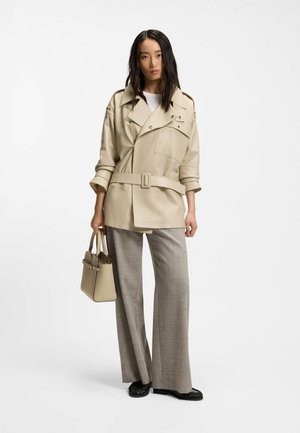 Nainen seisoo valkoista taustaa vasten, pukeutuneena beigenväriseen vyöllä varustettuun takkiin, leveälahkeisiin harmaisiin housuihin, mustiin kenkiin ja pitäen beigenväristä käsilaukkua.