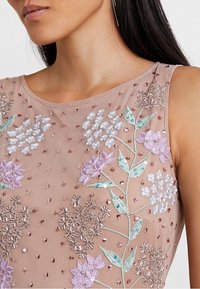 Top in tessuto beige con ricami floreali in viola, rosa e bianco, che presenta dettagli di paillettes e perline su una superficie testurizzata.