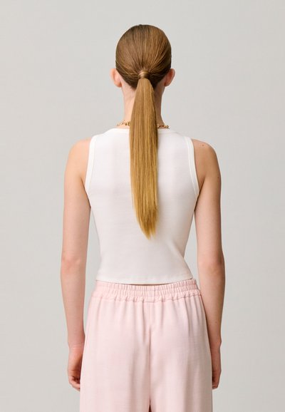 Mujer con el cabello largo y liso recogido en una coleta, vistiendo una camiseta blanca sin mangas y pantalones rosas con cintura elástica, de espaldas a la cámara.