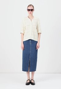 Hvit, strikket cardigan med knapper foran og korte ermer, kombinert med et langt, denimbunad med slisse foran, iført svarte leiligheter.