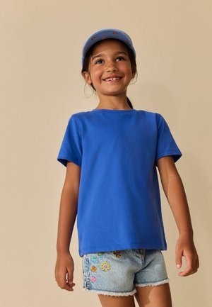 Naeratav tüdruk, kes kannab sinist mütsi ja t-särki ning lillemotiividega tikandiga teksapükse, taustaks lihtne beež sein.