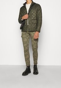 Quiltad olivgrön jacka med krage, ljusbeige tröja, kamouflagebyxor och svarta ankelskor. Axelremsväska.