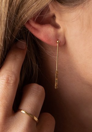 Boucles d'oreilles pendantes en or avec un clou en forme de boule arrondie et une barre rectangulaire texturée suspendue en dessous. Vue de près, portées avec des cheveux longs.