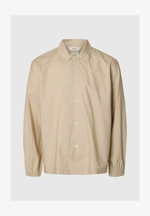 Beige pitkähihaiset nappilistallinen paita kuminauhahihoilla, leveällä kauluksella ja rento istuvuus, esillä yksivärisellä vaaleanharmaalla taustalla.