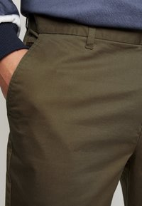 Zbliżenie na osobę noszącą oliwkowozielone spodnie, z ręką w przedniej kieszeni, w połączeniu z ciemną koszulką z długim rękawem z podwiniętymi mankietami.