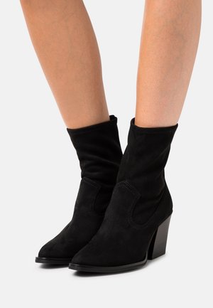 Czarne zamszowe botki za kostkę o luźnym fasonie, z wąskim czubkiem i słupkowym obcasem. Cechy obejmują detale ze szwów oraz miękką fakturę.
