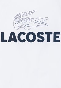 Tessuto bianco con un grande logo di coccodrillo nero sopra la parola "LACOSTE" in grassetto nero, con un motivo superficiale testurizzato.