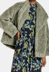 Chaqueta verde, texturizada y esponjosa con un cuello de chal, llevada sobre un vestido de estampado floral que presenta tonos azul oscuro, verde, amarillo y azul claro.