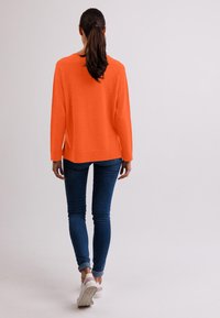 Maglione arancione lavorato a maglia con una vestibilità comoda, dotato di maniche lunghe e spacchi laterali, abbinato a jeans skinny blu scuro e sneakers bianche.