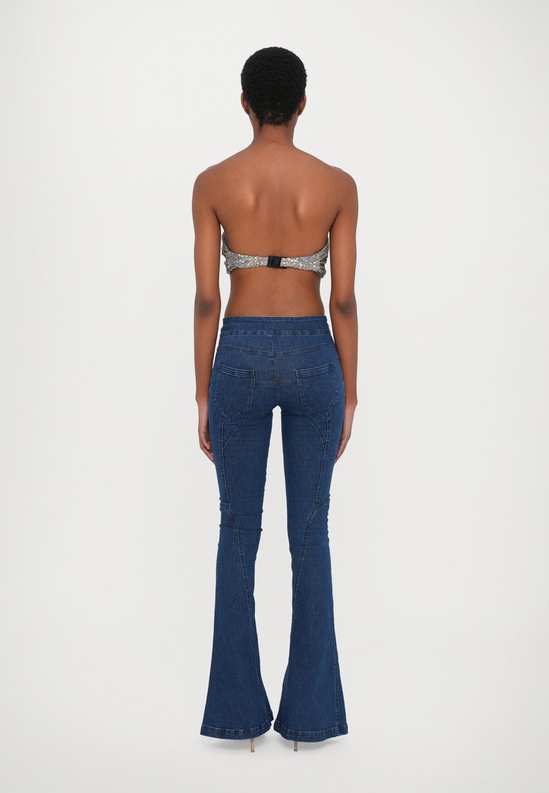 Bakifrån av en person som bär en grå paljetterad bralette och blå flarejeans med synliga sömnadsdetaljer och fickor. Platt midjelinje.