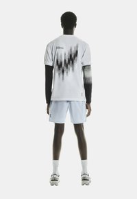 Witte sportt-shirt met zwart patroon op de achterkant, gecombineerd met lichtblauwe shorts. Model draagt witte sokken en grijze schoenen met zwarte accenten.