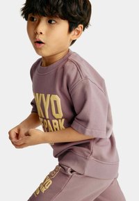 Sudadera lila con cuello redondo y mangas cortas, con texto amarillo en negrita. Combinada con pantalones jogger a juego, creando un conjunto cohesivo.