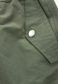 Nahaufnahme von olivgrünem Stoff mit einer gefalteten Lasche und einem einzelnen runden silbernen Druckknopf.