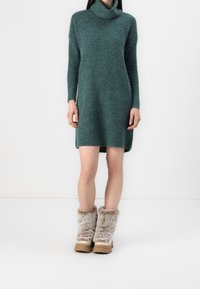 Robe en maille bleu sarcelle avec un col haut et des manches longues, associée à des bottes beiges duveteuses avec une apparence texturée et des semelles épaisses.