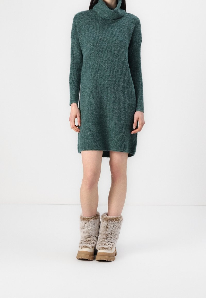 Robe en maille bleu sarcelle avec un col haut et des manches longues, associée à des bottes beiges duveteuses avec une apparence texturée et des semelles épaisses.