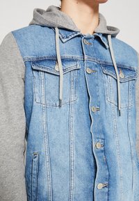 Jeansjacka med en ljusblå front och gråärmade. Har tryckknappar, två bröstfickor och en dragsko i huvan.