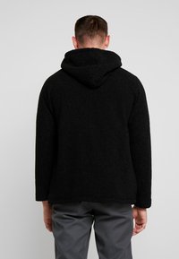 Sort, fuzzy hættetrøje med en struktureret ydre. Designet har en afslappet pasform med lange ærmer og en rund nederdel.