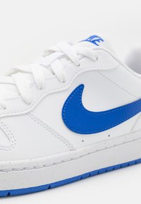 White sneaker with blue accents, featuring a smooth leather upper, textured side panel, stitched seams, and a rubber sole with blue tread.