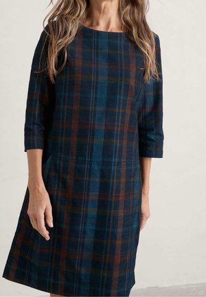 Kobieta w sukience w kratę o długości do kolan z rękawami do połowy przedramienia w odcieniach niebieskiego, brązowego i zielonego na jednolitym tle.