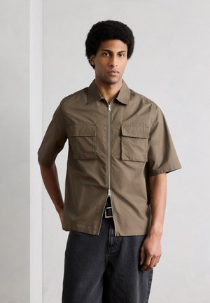 Man with afro hairstyle wearing a short-sleeve khaki zip-front shirt with chest pockets and black pants, standing against a plain wall.