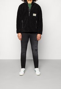 Sudadera negra y peluda con cierre frontal y bolsillos laterales; llevada sobre una blusa verde, combinada con jeans oscuros y zapatillas blancas.