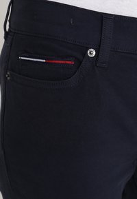 Pantalones de algodón azul marino con un pequeño bolsillo frontal con franjas en rojo, blanco y azul, y un detalle de botón plateado en la cintura.