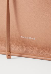 Svetlo rjav usnjen nahrbtnik z teksturirano površino. Ima minimalističen dizajn in logotip "Coccinelle" v beli barvi, ki je postavljen v spodnjem delu.