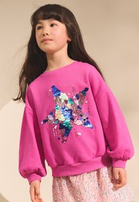 Roza jopa iz mehke tkanine z večbarvnim vzorcem zvezde iz sequins. Rokavi so napihnjeni in z rebrastimi manšetami na zapestjih in spodnjem robu.