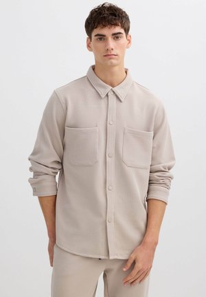 Camisa beige claro abotonada, hecha de tela texturizada, con cuello, dos bolsillos en el pecho y mangas remangadas.