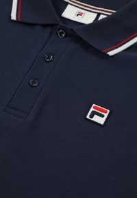 Polo bleu marine avec trois boutons noirs, col côtelé avec rayures blanches et rouges, et patch logo Fila sur la poitrine.