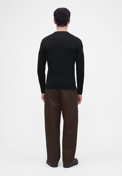 Homme de dos portant un pull noir ajusté à manches longues, un pantalon marron ample et des chaussures noires sur un fond blanc uni.